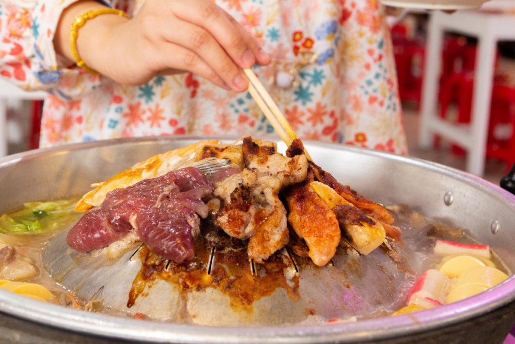 A girl at home is holding chopsticks over a hotplate of food delivered in a mookata set.